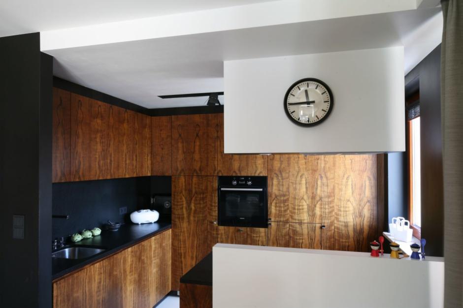 Nowoczesna kuchnia z drewnianymi szafkami o wyraźnym usłojeniu, czarnym blatem i elementami w kolorze czarnym i białym. Na ścianie zegar.