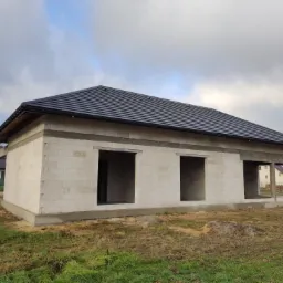 Surowy, jednopiętrowy budynek z białych bloczków z zamontowanym czarnym dachem, widoczne otwory okienne i drzwiowe, w tle inne domy i pochmurne niebo.