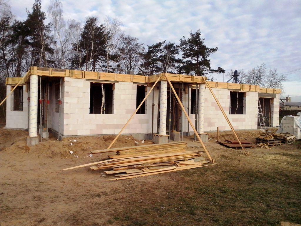 Niedokończony parterowy budynek z betonu komórkowego, drewniane szalunki nad otworami okiennymi i słupami, podparte drewnianymi stemplami, widoczne deski i drabina, jesienne drzewa w tle.