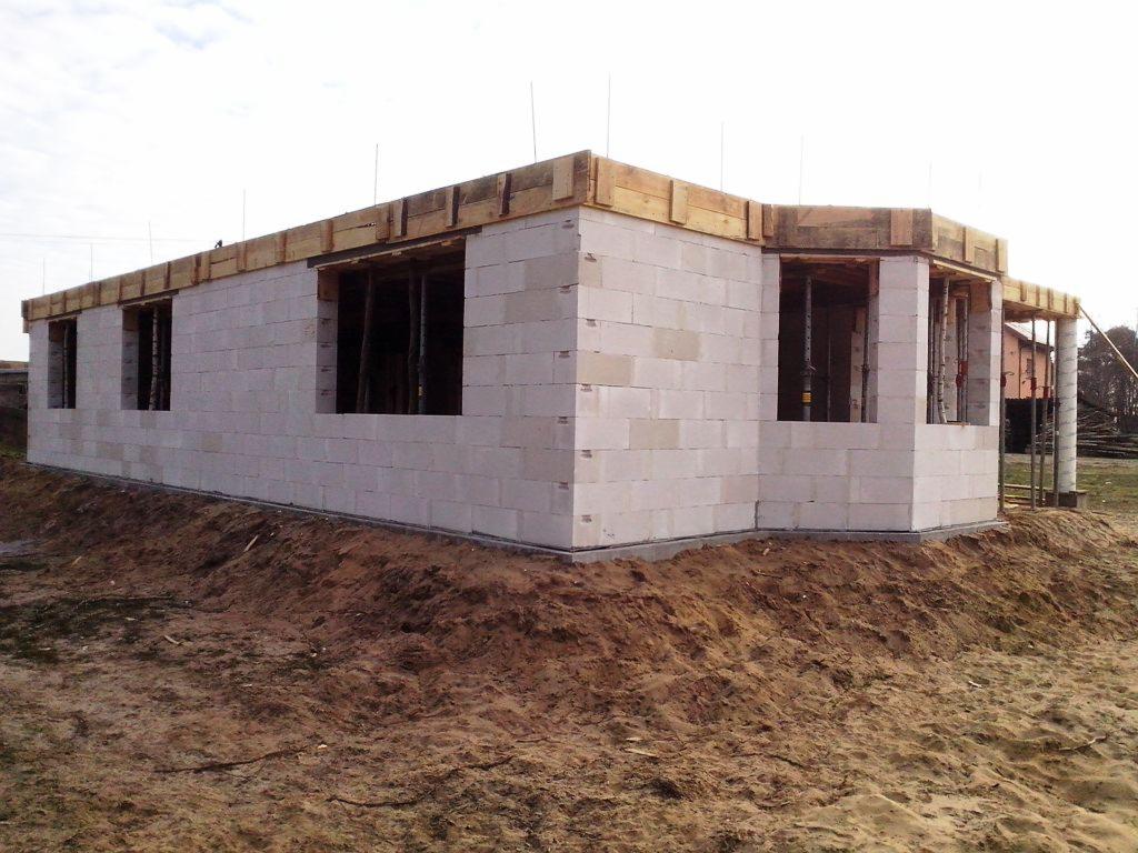 Surowy stan domu z betonu komórkowego z widocznymi otworami okiennymi i drewnianym szalunkiem wieńczącym ściany, otoczony ziemią.