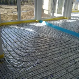 Białe rury ogrzewania podłogowego ułożone spiralnie na folii z nadrukiem siatki, widoczne okna z widokiem na plac budowy i żółta folia ochronna przy ścianach.