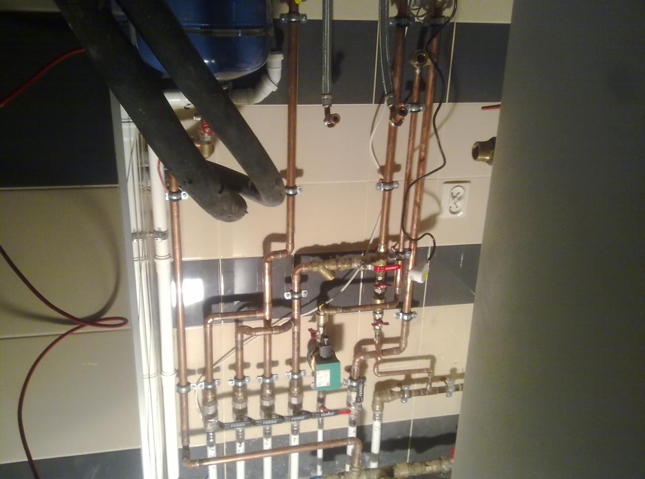 Złożona instalacja hydrauliczna z miedzianych rur, zaworów i przewodów, zamontowana przy ścianie wyłożonej biało-szarymi płytkami, widoczny fragment bojlera oraz izolowane rury.