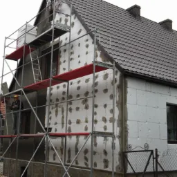 Dom w trakcie ocieplania: styropian z zaprawą, rusztowania, pracownik w kasku. Częściowo otynkowana ściana, brązowy dach. Jesienne drzewa w tle.