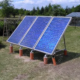 Trzy kolektory słoneczne z niebieskimi panelami na trawniku, wsparte na pomarańczowych rurach, w tle zielony ogród i szklarnia. Ujęcie z niskiej perspektywy.