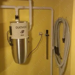 Centralny odkurzacz Duovac 45 Star zamontowany na żółtej ścianie w Bielsko-Białej. Widoczne rury instalacji i wąż ssący.