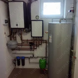 Instalacja kotła gazowego z zasobnikiem ciepłej wody i filtrami wody, widoczne miedziane rury i osprzęt, umieszczone w pomieszczeniu technicznym z oknem.