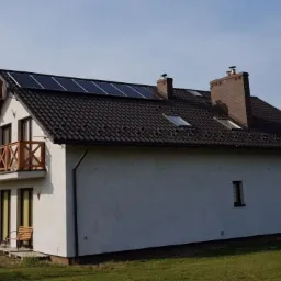 Dom jednorodzinny z ciemną dachówką i panelami fotowoltaicznymi na dachu, widoczny balkon z drewnianą balustradą i powiewająca polska flaga.