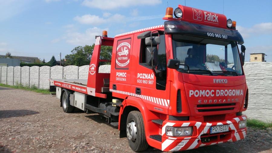Czerwona laweta marki Iveco z napisem 'Pomoc Drogowa Poznań' i numerem telefonu, zaparkowana na żwirowym podłożu z betonowym ogrodzeniem w tle.