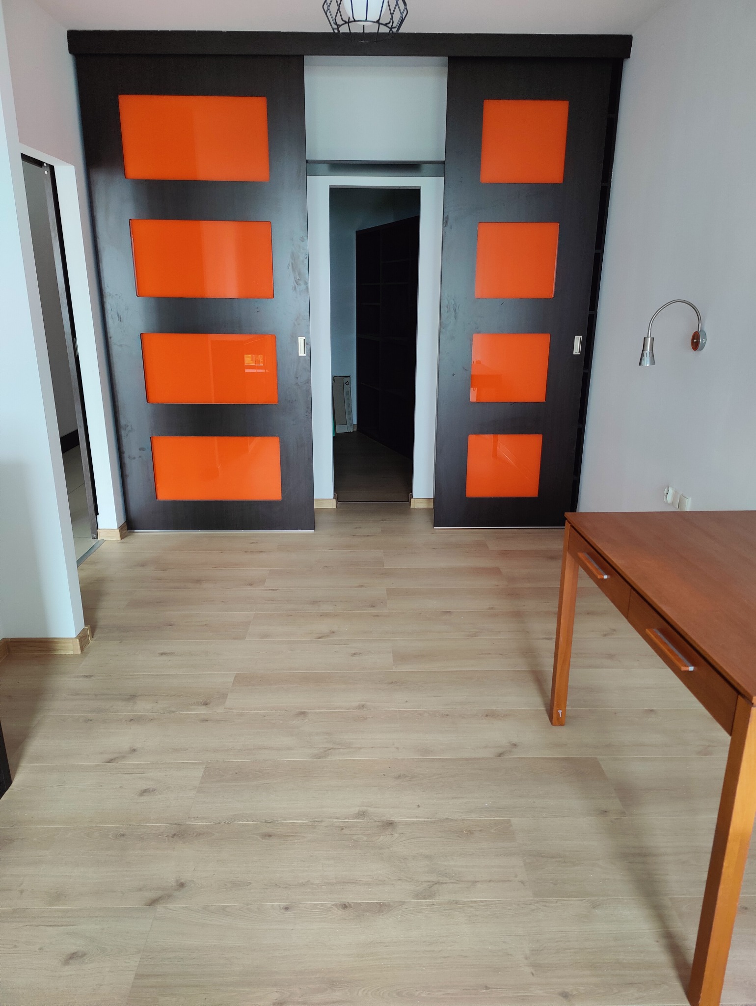 Jasne panele podłogowe i przesuwne drzwi z pomarańczowymi wstawkami w Piasecznie. Widoczny fragment drewnianego stołu i lampka na ścianie. Nowoczesne wykończenie wnętrza.