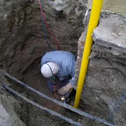 Pracownik w wykopie spawa rury, widoczna ż&oacute;łta rura osłonowa i narzędzia. Prace ziemne przyłącza gazowego z użyciem spawarki.