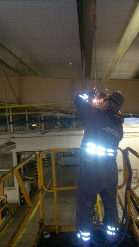 Spawacz w kombinezonie roboczym, w okularach ochronnych, spawa rurę na wysokości na tle hali przemysłowej. Widoczne iskry i elementy konstrukcyjne.