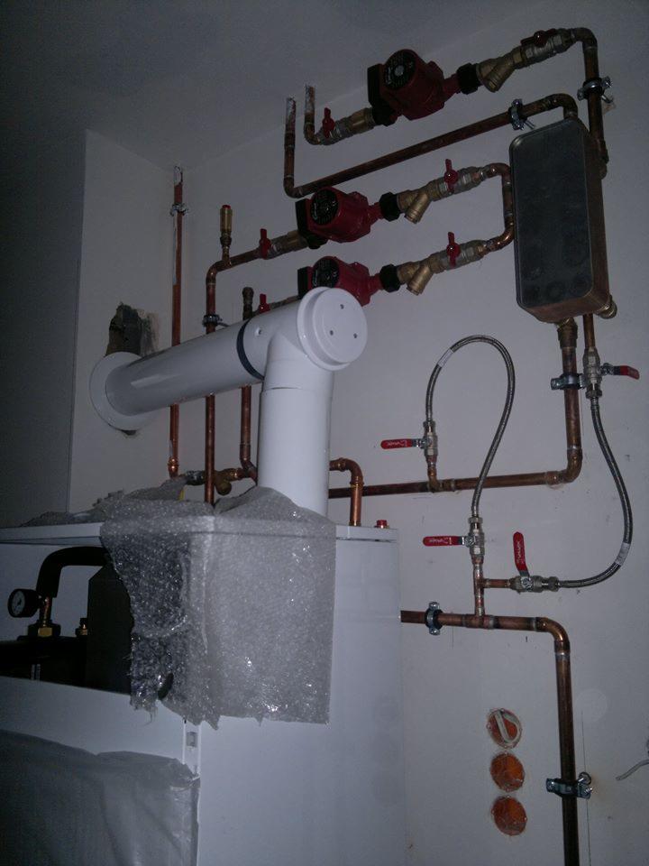 Złożona instalacja grzewcza z miedzianymi rurami, pompami obiegowymi i białym zasobnikiem. Część zasobnika zabezpieczona folią bąbelkową. Widoczne zawory z czerwonymi pokrętłami.