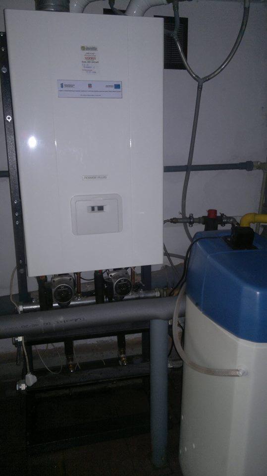 Biały piec gazowy Beretta Power Plus zamontowany na ścianie, widoczne rury, pompy obiegowe i filtr wody. Ujęcie w pomieszczeniu technicznym.