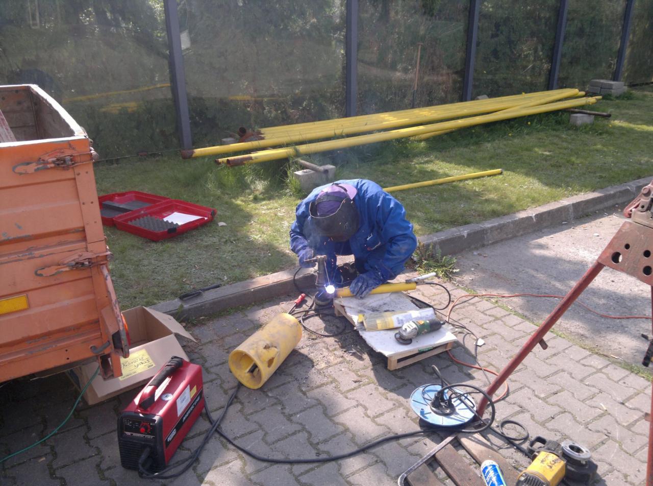 Spawanie żółtej rury gazowej przez spawacza w niebieskim kombinezonie na zewnątrz, widoczne narzędzia i materiały, trawa w tle.