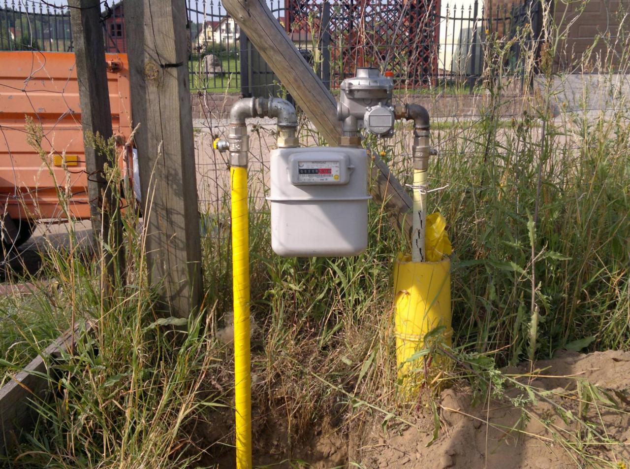 Szary gazomierz z żółtą rurą w ziemi, połączony metalowymi rurami. Widoczne trawy i ogrodzenie w tle. Realizacja przyłącza gazowego.