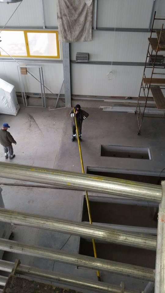 Dwóch pracowników w hali produkcyjnej, jeden mierzy żółtą rurę w otworze w podłodze, drugi stoi obok. Widok z góry na instalację i drabiny.