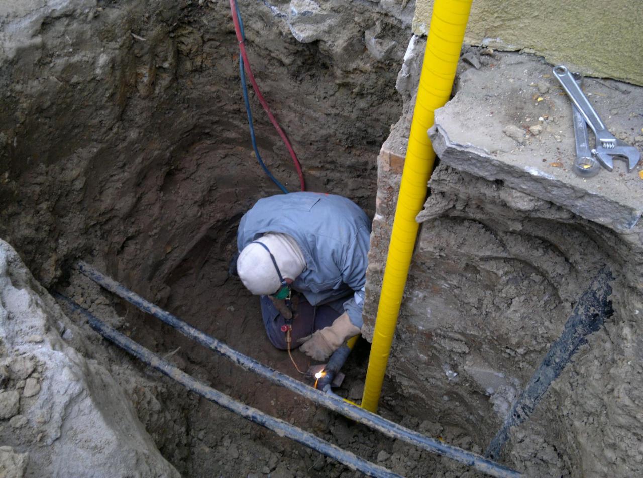 Pracownik w wykopie spawa rury, widoczna żółta rura osłonowa i narzędzia. Prace ziemne przyłącza gazowego z użyciem spawarki.