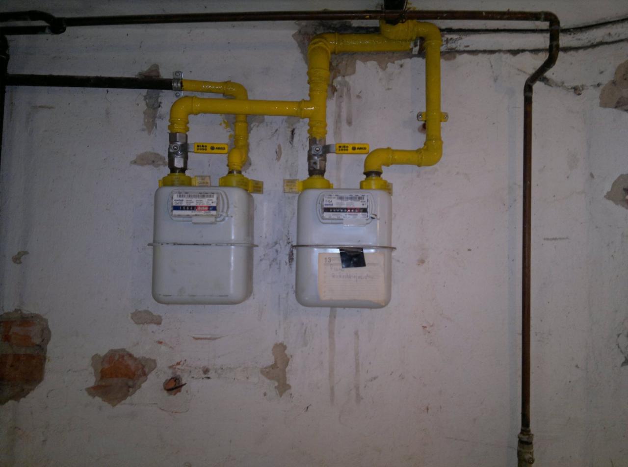 Dwa gazomierze z żółtą instalacją gazową na tle ściany z widocznymi fragmentami cegieł. Rury biegną wzdłuż ściany. Widoczne zawory odcinające dopływ gazu.
