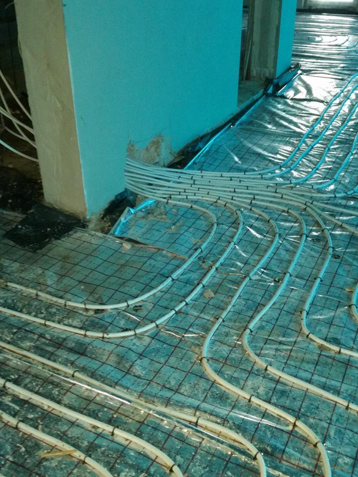 Białe rurki ogrzewania podłogowego ułożone na siatce z folią izolacyjną, widoczne fragmenty ściany w surowym stanie, przygotowane do zalania wylewką.