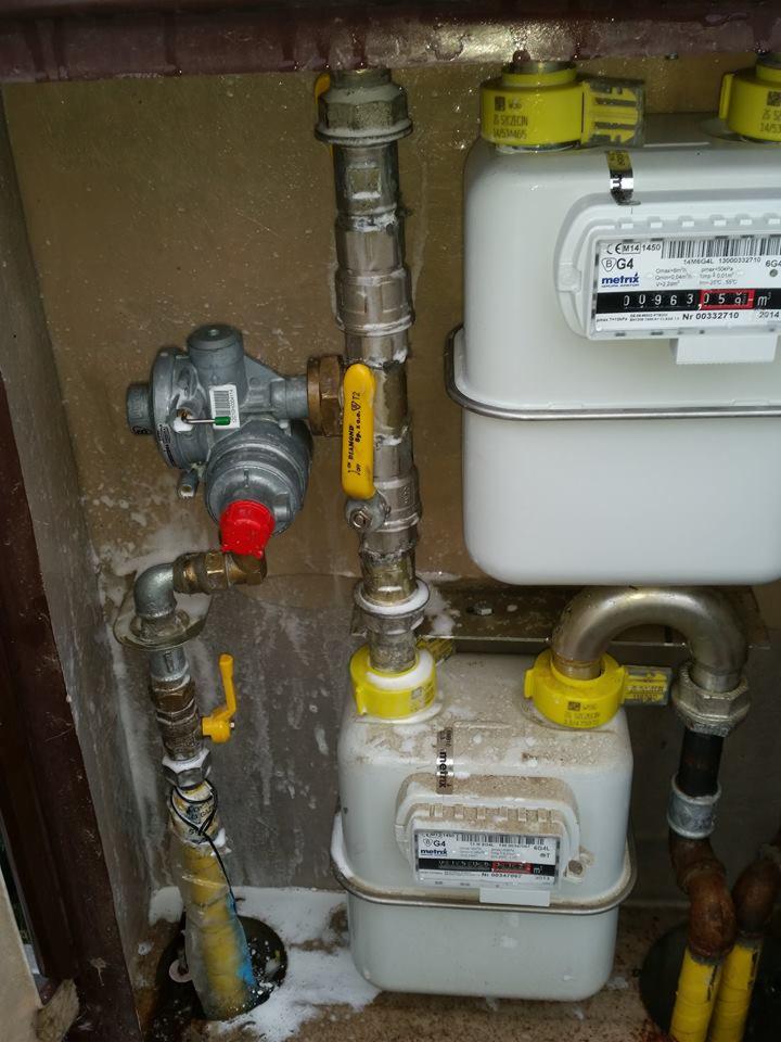 Kompleksowy montaż gazomierza z reduktorem i zaworami, rury stalowe z żółtymi elementami, instalacja wewnątrz budynku, białe ślady uszczelnienia na połączeniach.