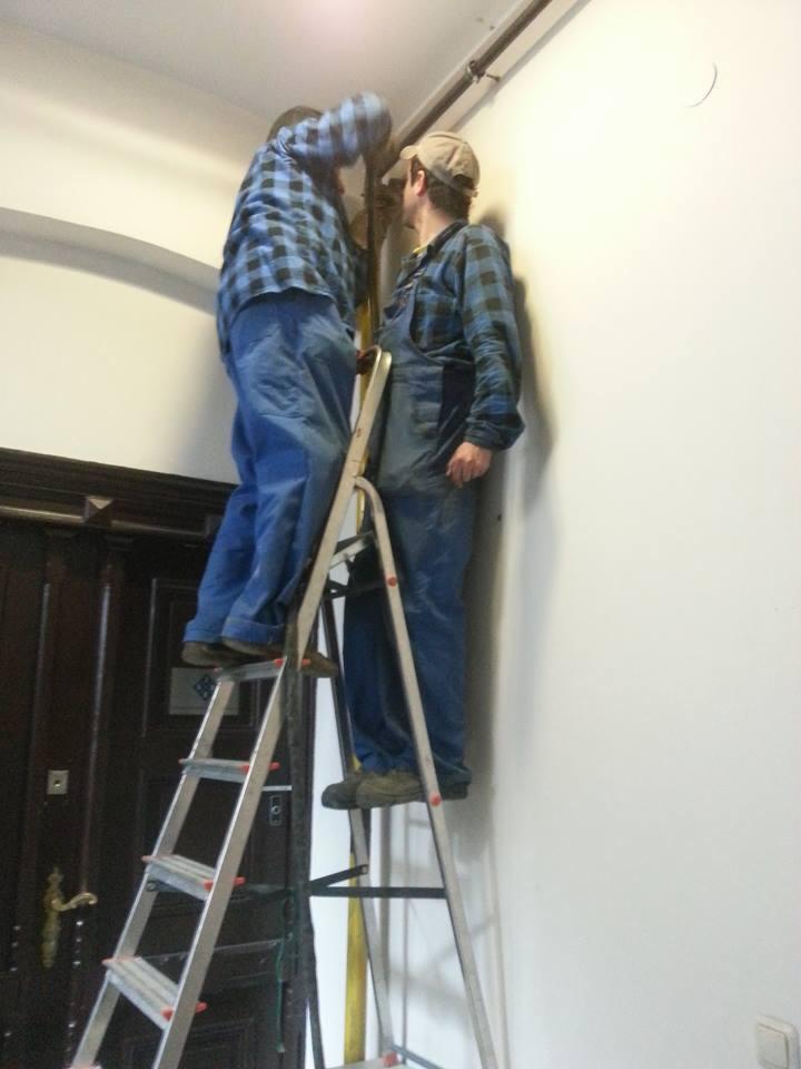 Dwóch monterów na drabinie, w niebieskich kombinezonach, instaluje rurę pod sufitem w jasnym pomieszczeniu. Widoczna stara, ciemna stolarka drzwiowa.