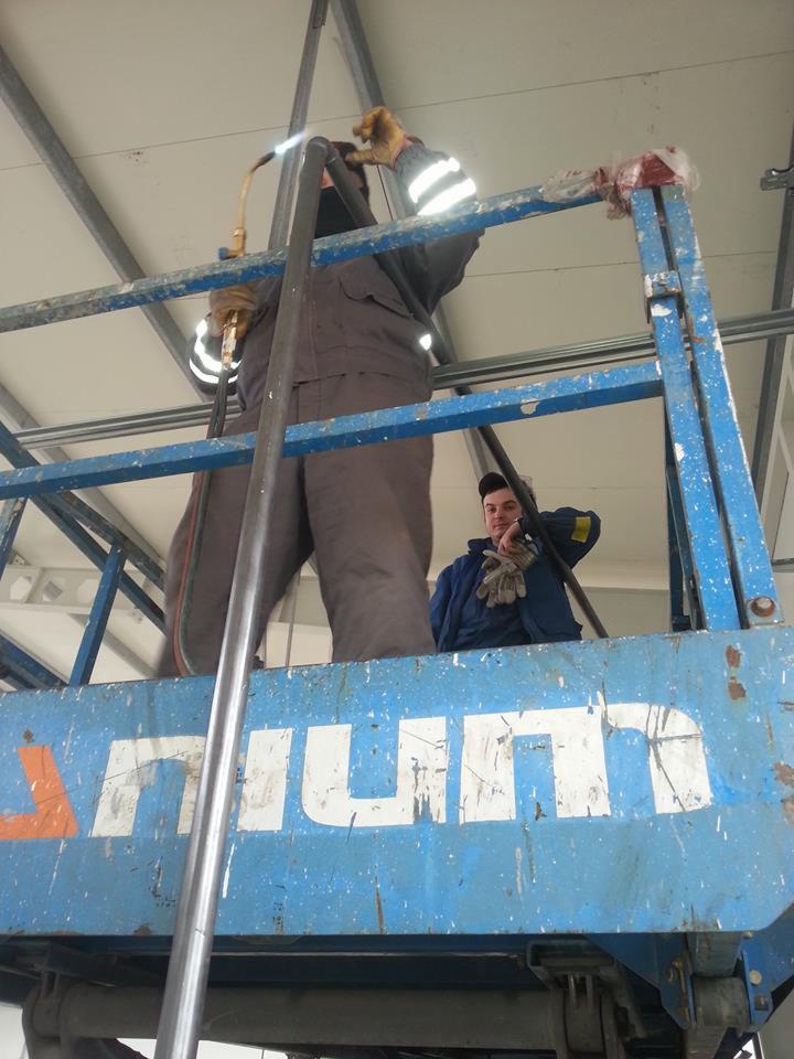 Pracownik na podnośniku spawa rurę palnikiem, drugi obserwuje, widoczne elementy konstrukcji sufitu. Praca na wysokości, odzież robocza.