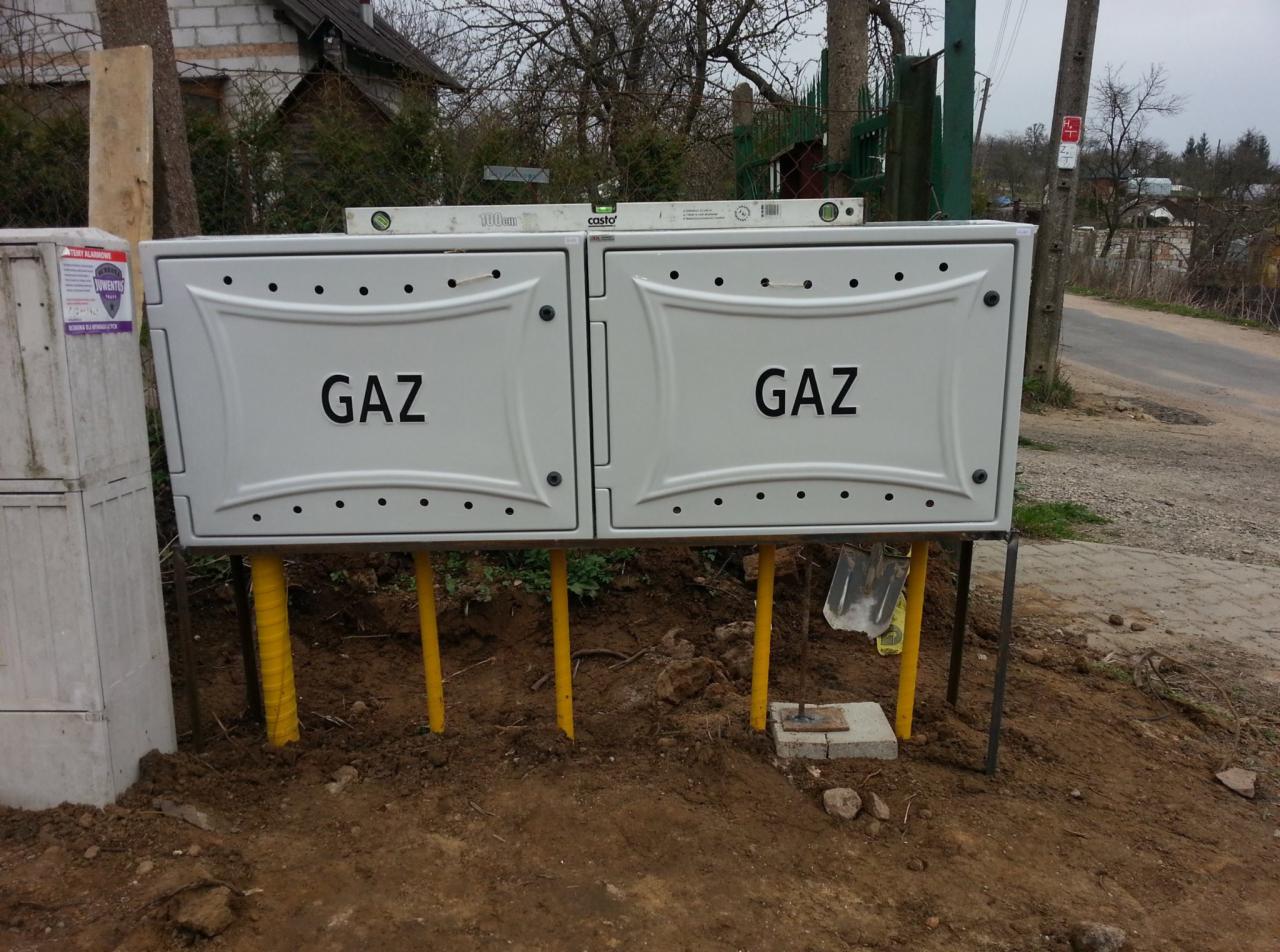 Dwa białe, symetryczne gazowe przyłącza, każde z napisem 'GAZ', zamontowane na żółtych rurach. Poziomica na górze. Widoczne narzędzia i teren po instalacji.