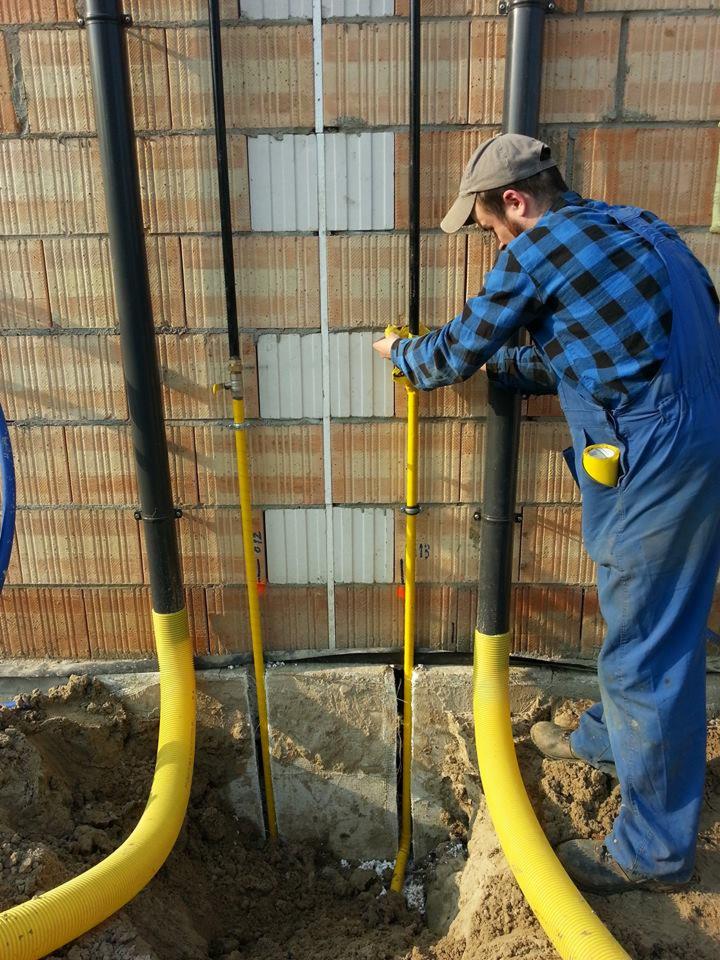 Instalator w niebieskim kombinezonie łączy żółte rury gazowe do ściany z cegieł. Widoczne czarne rury i wykopane fundamenty. Taśma miernicza w dłoniach.