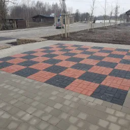 Ułożona kostka brukowa w szachownicę z czerwonych i grafitowych kostek, otoczona szarą kostką brukową, na tle parkingu i placu zabaw.