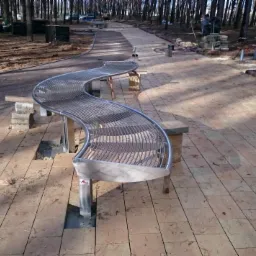 Falista ławka ze stali nierdzewnej na tle parku z brukowaną ścieżką. W tle widoczne drzewa i pracownik układający kostkę.