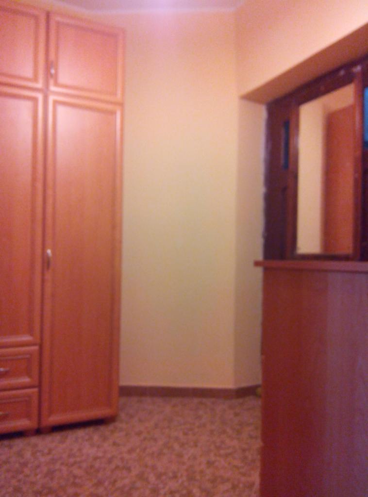 Pokój po remoncie: brązowa szafa z szufladami, beżowe ściany, wzorzysta podłoga i zabudowa okienna z brązowego drewna. Widok na odnowione wnętrze.