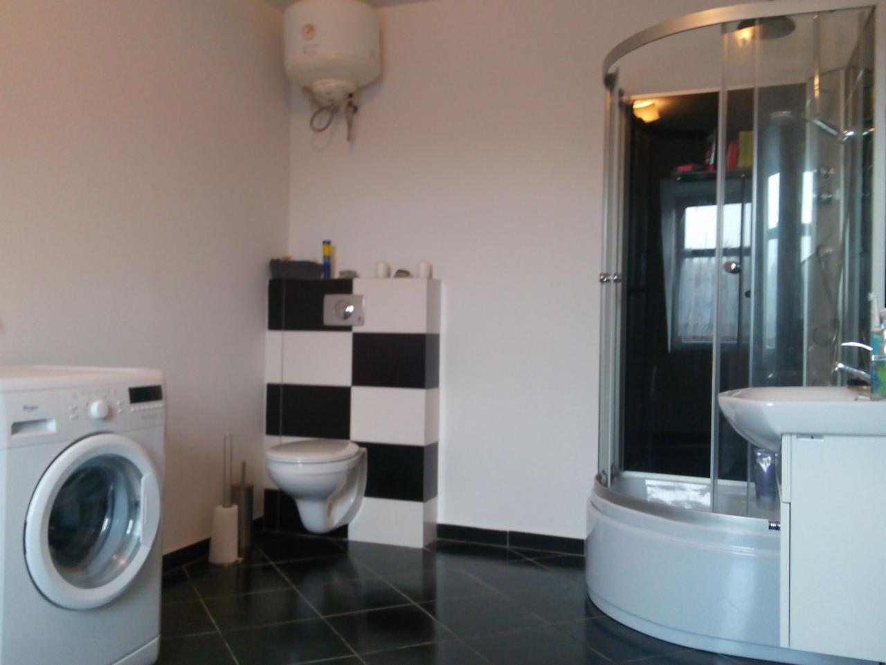 Nowoczesna łazienka z kabiną prysznicową, pralką i toaletą w czarno-białej szachownicy. Minimalistyczny design, czarne płytki podłogowe, bojler na ścianie.