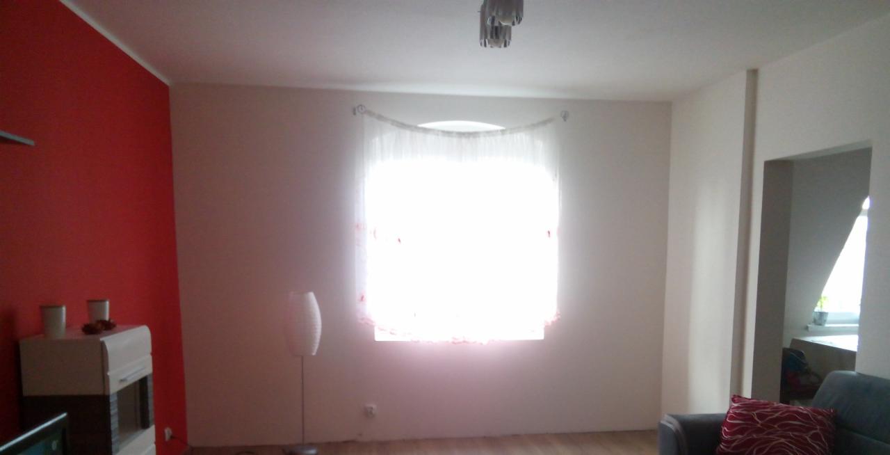 Salon po odświeżeniu: czerwona ściana, jasne ściany, okno z firanką. Widoczny fragment sofy i komody. Podłoga drewniana. Lampa podłogowa.