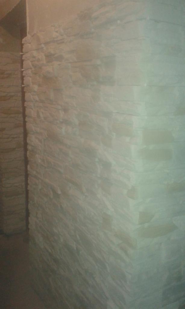 Ściana wykończona okładziną z jasnego kamienia dekoracyjnego, imitującego naturalny kamień, z widocznym narożnikiem i fragmentem sąsiedniej ściany.