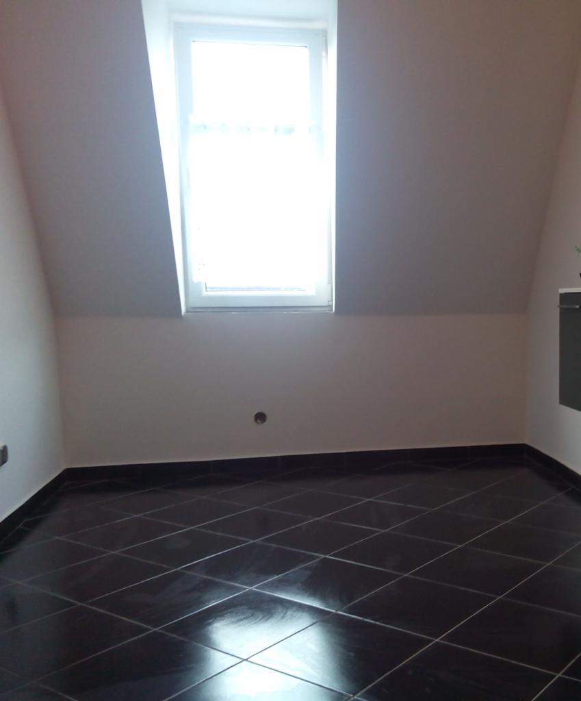 Pokój na poddaszu z oknem i czarną, błyszczącą podłogą z płytek ułożonych w karo. Ściany białe, gładkie. Widoczny fragment szafki po prawej stronie.