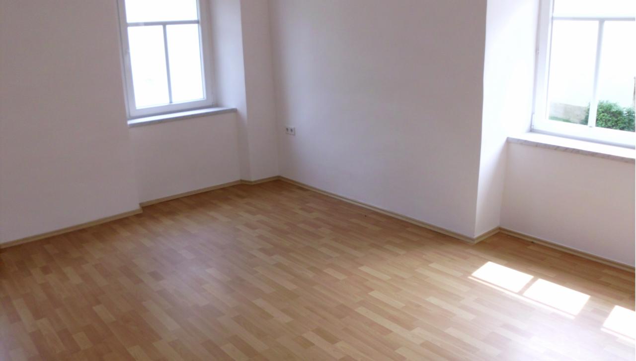 Jasne, puste wnętrze z drewnianą podłogą, dwoma oknami z marmurowymi parapetami i białymi ścianami. Widoczne gniazdko elektryczne. Promienie słoneczne na podłodze.