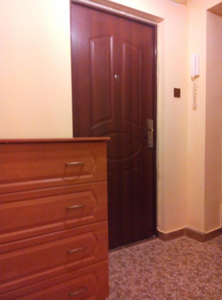 Wnętrze mieszkania z brązowymi drzwiami, komodą i domofonem na ścianie. Podłoga wyłożona terakotą. Ciepłe oświetlenie.