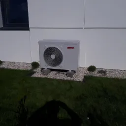 Zewnętrzna jednostka pompy ciepła Ariston zamontowana na betonowych podstawach przy białej ścianie budynku, otoczona białym żwirem i trawnikiem.