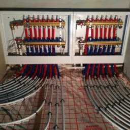 Rozdzielacz ogrzewania podłogowego w białej szafce z licznymi czerwonymi i niebieskimi rurkami, ułożony na folii z czerwonym nadrukiem siatki.