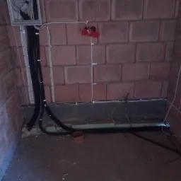 Surowa ściana z czerwonej cegły z widoczną instalacją elektryczną w trakcie montażu, obejmującą czarne i białe kable oraz puszkę rozdzielczą.