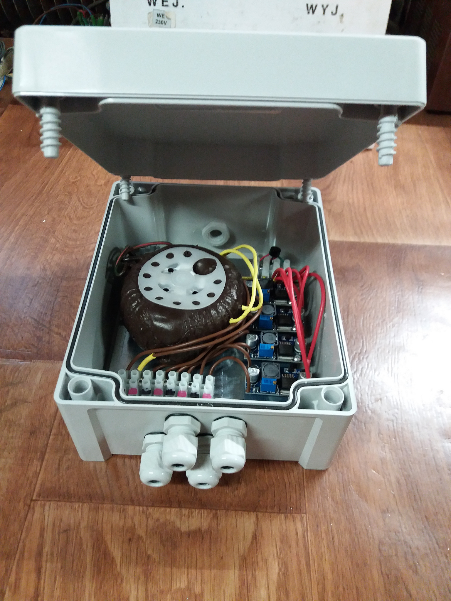 Otwarta szara skrzynka elektryczna z transformatorem toroidalnym, modułami zasilania i złączkami kablowymi na drewnianej podłodze.