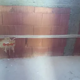 Wnętrze w trakcie prac: kable elektryczne ułożone na ceglanej ścianie z wyciętymi otworami pod gniazdka, widoczne fragmenty betonu. Surowy stan, bez wykończenia.