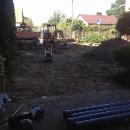 Żółta koparka i zagęszczarka na placu budowy z wykopaną ziemią, w tle ciężarówka i rury kanalizacyjne na pierwszym planie.