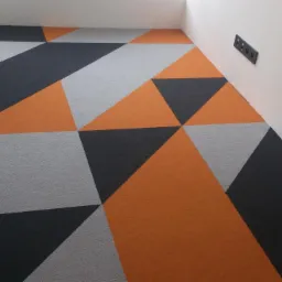 Wzorzysta wykładzina podłogowa w odcieniach szarości, czerni i pomarańczy, ułożona w geometryczny deseń z trójkątów i kwadratów. Widoczne gniazdka elektryczne na ścianie.