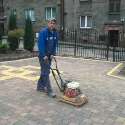 Pracownik zagęszcza kostkę brukową z żółtymi wstawkami na podjeździe przed budynkiem. W tle drugi pracownik i roślinność.