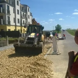 Żółta koparko-ładowarka CAT na tle budynku mieszkalnego i ekipy budowlanej podczas utwardzania drogi kruszywem. Widoczny pracownik w kamizelce odblaskowej.