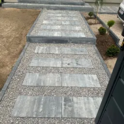 Nowoczesna ścieżka z płyt betonowych i grysu prowadząca do domu z niebieskimi drzwiami. Estetyczne wykończenie ogrodu z roślinami wzdłuż ścieżki.