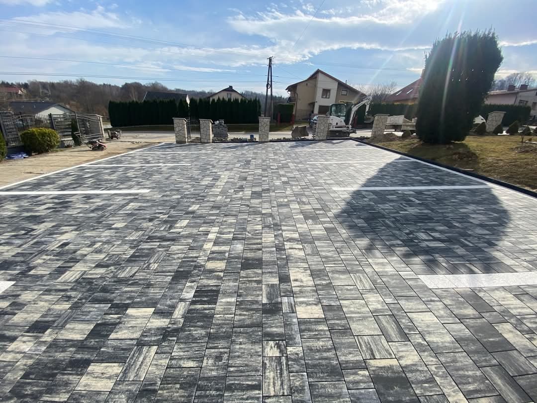 Ukończony parking z kostki brukowej w odcieniach szarości, wyznaczony białymi liniami, z mini koparką w tle i elementami ogrodzenia w oddali.