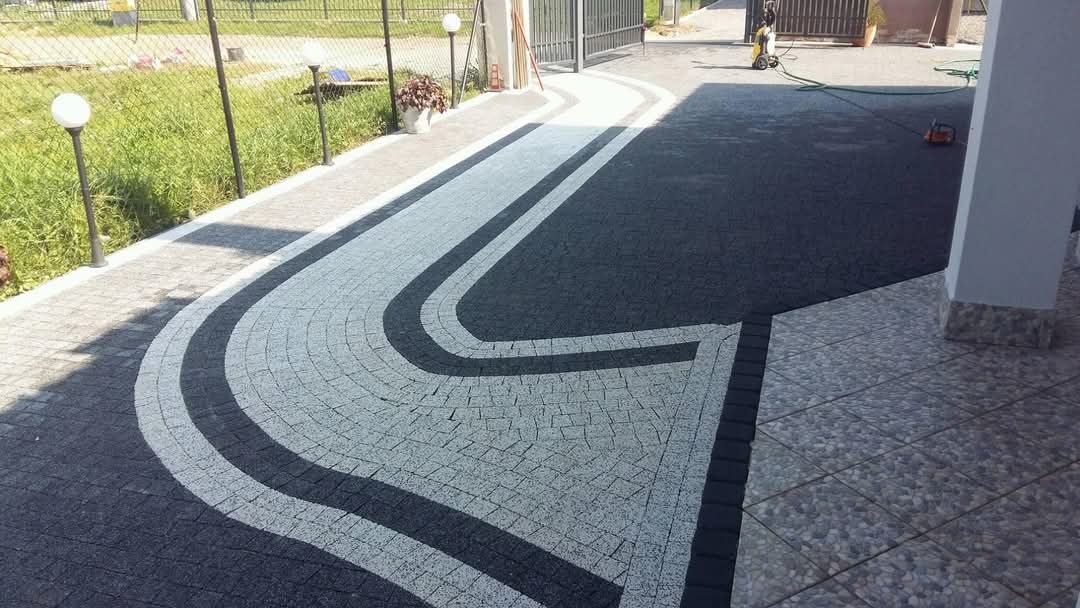 Wzorzysta nawierzchnia z kostki granitowej w odcieniach szarości i czerni, tworząca elegancki podjazd przed domem z widocznymi lampami ogrodowymi.