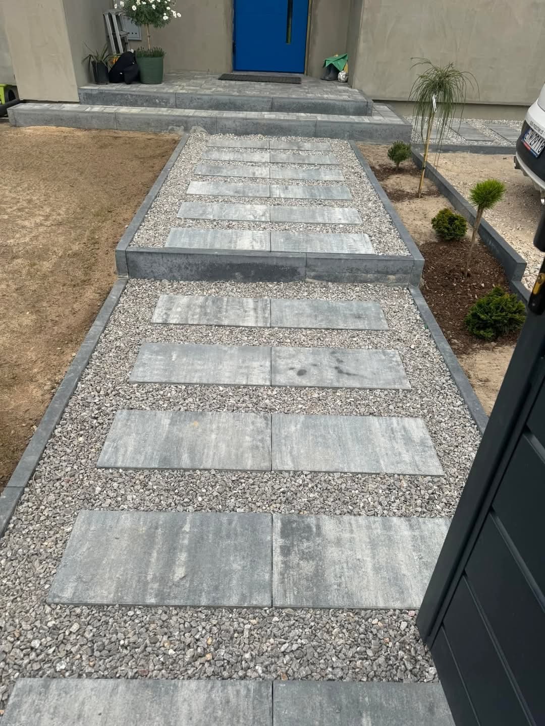 Nowoczesna ścieżka z płyt betonowych i grysu prowadząca do domu z niebieskimi drzwiami. Estetyczne wykończenie ogrodu z roślinami wzdłuż ścieżki.